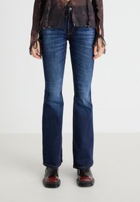 Vaqueros acampanados de mezclilla en azul oscuro con una textura suave, un ligero desvanecimiento y costuras en contraste. Combinados con botas brillantes negras y burdeos.