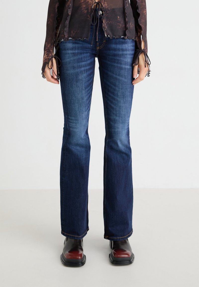 Vaqueros acampanados de mezclilla en azul oscuro con una textura suave, un ligero desvanecimiento y costuras en contraste. Combinados con botas brillantes negras y burdeos.