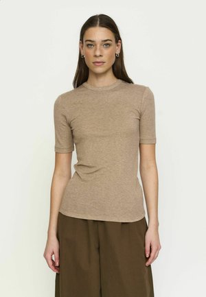 Woman with straight brown hair wears a fitted beige short-sleeve top and wide-leg brown pants, standing against a plain light background.
