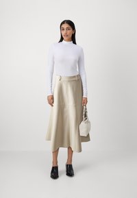 Haut blanc à manches longues, jupe midi en cuir beige avec des détails de couture, associée à des mules noires et un petit sac blanc avec une bandoulière texturée.