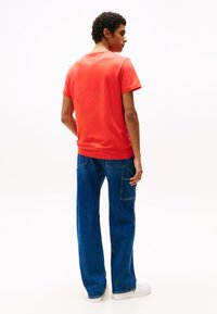 Tricou roșu cu guler rotund asociat cu blugi lejeri de denim albastri. Caracteristici notabile includ o croială dreaptă și un buzunar lateral pe blugi.