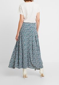 Floraler Maxirock in Blau, Grün und Weiß mit Falten; kombiniert mit einem schlichten weißen T-Shirt und cremefarbenen Stiefeln mit Absatz, die den fließenden Stoff zur Geltung bringen.