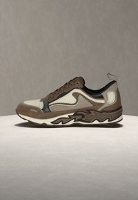 Καφέ και μπεζ αθλητικό παπούτσι με πάνελ από δίχτυ, σουέντ λεπτομέρειες, παχύ σόλα με σχέδιο, δέσιμο με κορδόνια και ταινία στο πίσω μέρος, τοποθετημένο σε ουδέτερη επιφάνεια.