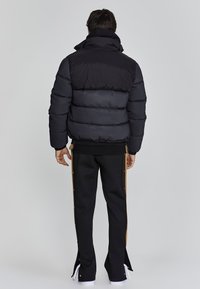 Schwarze Steppjacke mit hochgeschlossenem Kragen und gestepptem Design. Kombiniert mit schwarzen Hosen mit seitlichen Druckknöpfen und hellbraunen Akzenten.