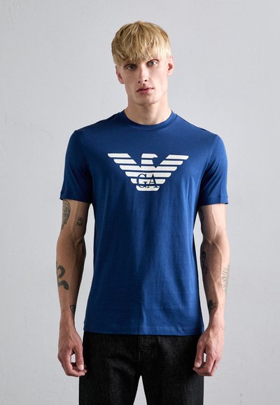 Camiseta de algodón azul marino con un gráfico de águila blanca y rayas. Corte ajustado con mangas cortas, sobre un fondo gris claro.