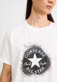 Camiseta de algodón blanca con un gráfico circular negro con una estrella en el centro, texto "CONVERSE," "Chuck," y "ALL STAR," con acentos de salpicaduras.