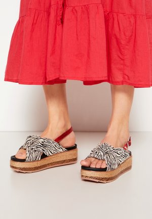 Füße mit Plateausandalen mit Zebra-Print-Riemen und roten Knöchelriemen, kombiniert mit einem roten Midirock.