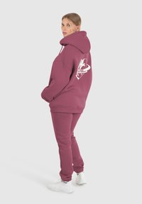 Hoodie oversize color marsala con tasche frontali, polsini a coste e una stampa grafica sul retro, abbinato a pantaloni della tuta coordinati e sneakers bianche.