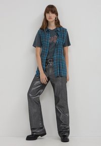 Camicia a quadri turchese sovrapposta a una maglietta grafica, abbinata a pantaloni larghi in argento metallico e stivali chunky neri. Gli accessori includono anelli.