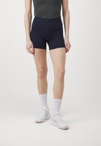 Σκούρα μπλε ψηλόμεσα σορτς από ελαστικό υλικό, με φαρδύ λάστιχο στη μέση και μήκος που φτάνει μέχρι το μέσο του μηρού, συνδυασμένα με λευκά αθλητικά παπούτσια και κάλτσες.