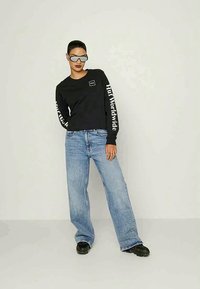 Oseba v črni majici z dolgimi rokavi, na katerih je napis "Huf Worldwide", v širokih modrih kavbojkah, črnih čevljih in z odbojnimi sončnimi očali.