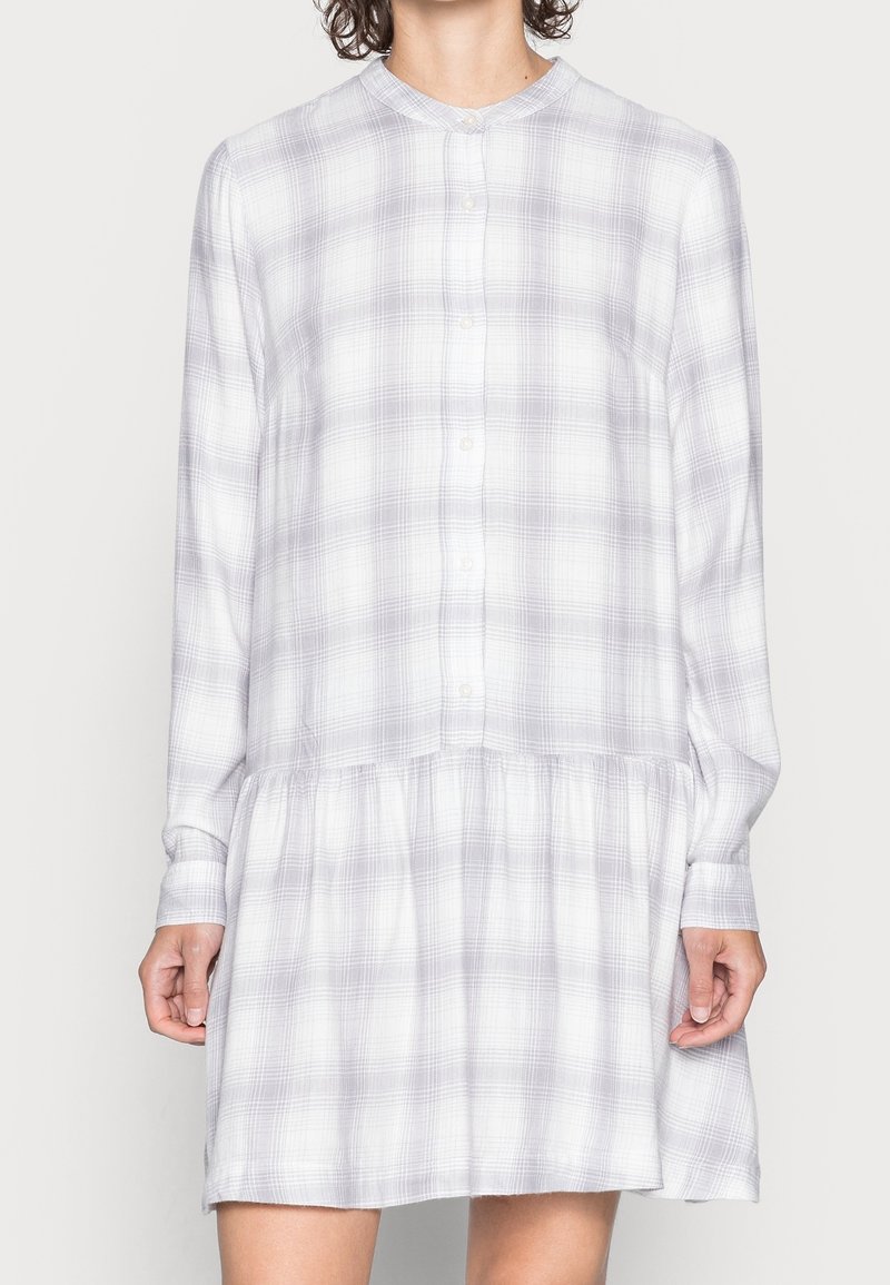 Femme portant une robe longue à manches longues, boutonnée, en gris clair à carreaux, avec une taille froncée et un col rond.