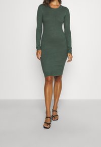 Abito a maniche lunghe in maglia a costine verde scuro, silhouette aderente, scollo rotondo, che arriva a metà coscia, abbinato a sandali neri con cinturini.