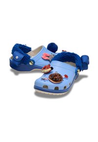 Helleblaue Clogs mit blauer, flauschiger Innenauskleidung, verziert mit bunten Charms von Cartoonfiguren und Lebensmitteln auf weißem Hintergrund.