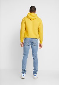 Gul hoodie med dragsko, avslappnad passform, kombinerad med ljusblå jeans och vita sneakers med blå accenter. Enfärgad bakgrund.