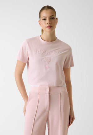 Vrouw draagt een lichtroze GUESS cropped T-shirt en getextureerde, hooggetailleerde roze broek, staand tegen een effen lichte achtergrond.