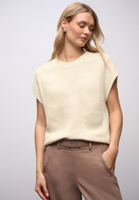 Haut en maille beige clair à manches courtes, présentant un motif texturé et un col rond, associé à un pantalon marron et une ceinture.