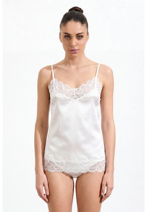 Woman with hair in bun wearing white satin camisole and matching lace-trimmed underwear, standing against a plain white background.
