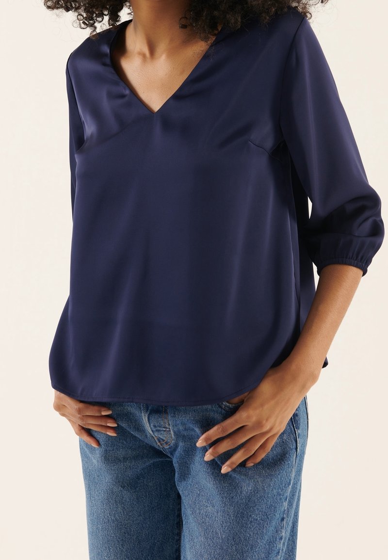 Femme portant un chemisier bleu marine à manches longues avec un col en V et des poignets élastiques, ainsi qu'un jean en denim bleu, les mains posées sur les hanches.