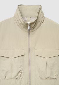 Beige leichte Jacke mit hohem Kragen, durchgehendem Reißverschluss vorne und zwei großen Brusttaschen mit Falten. Glatte, leicht geknitterte Textur.