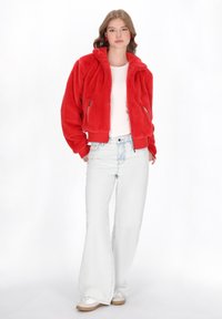 Jachetă bomber din blană artificială roșie, cu fermoar frontal, margine ribbed, buzunare laterale cu fermoar, purtată peste o cămașă albă și blugi largi de culoare albastră deschisă.