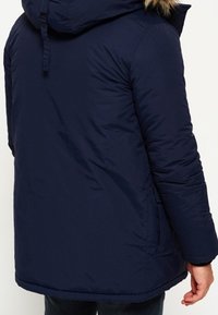 Chaqueta acolchada azul marino con capucha, con ribete de piel sintética, textura acolchada y bolsillos laterales. El tejido parece resistente al agua.