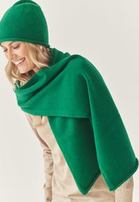 Grøn uldhat og sjal, blød tekstur, draperet design. Sjalet har glatte kanter, båret over en beige top. Naturligt lys.