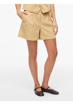 Kobieta w beżowych plisowanych szortach z bocznymi kieszeniami i czarnych balerinach typu Mary Jane, stojąca na białym tle.