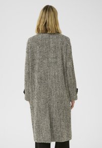 Personne avec des cheveux blonds arrivant aux épaules, portant un long manteau en herringbone noir et blanc, se tenant devant un fond clair.