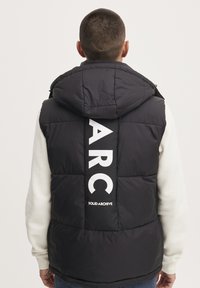 Schwarze gepolsterte Weste mit Kapuze, die auf der Rückseite große weiße Buchstaben "ARC" und "SOLID ARCHIVE" aufgedruckt hat. Glatte Textur, warmes Design.