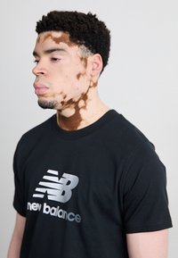 T-shirt nera con un grande logo riflettente argento "New Balance" sul davanti. Il modello ha i capelli corti e ricci e un tono di pelle chiaro.