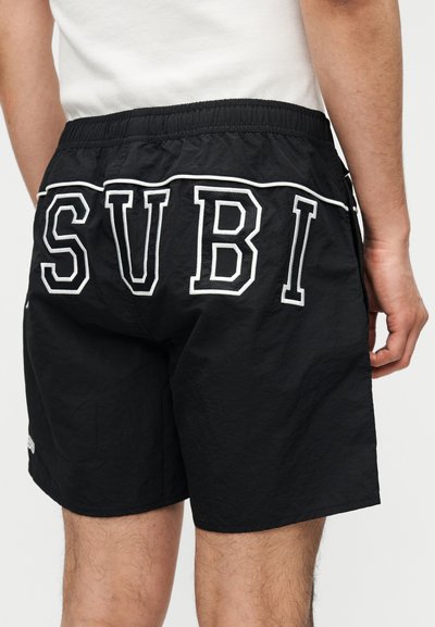 Vyras dėvi juodus trumpus šortus su elastinga juosmens juosta ir didelėmis balto raidėmis „SUBI“ ant nugaros, derindamas su baltu marškinėliu.