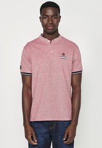 Raudona trumpomis rankovėmis polo marškinėliai su henley tipo apykakle, su mažuoju logotipu, dryžuota krūtinės kišenė ir kontrastingais juodais akcentais ant rankovių.