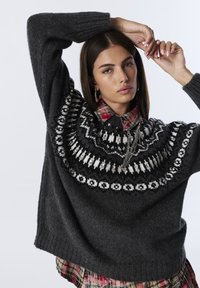 Maglione oversized grigio scuro con un giogo decorato in bianco e nero, caratterizzato da un collo alto e polsini a costine, indossato sopra una camicia scozzese.