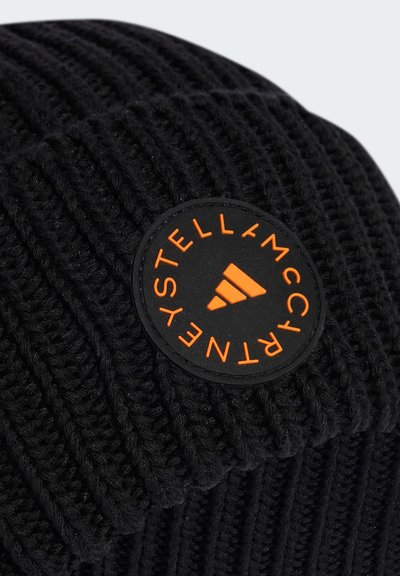 Melna adīta cepure ar vertikālu ribbingu, ar apaļu melnu uzlīmi, uz kuras redzams oranžs teksts un logo. Mīksta tekstūra un ikdienišķs dizains.