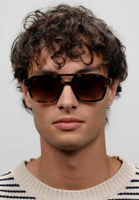 Occhiali da sole tartarugati con lenti scure, design della montatura geometrico e incassi in metallo. Il modello indossa un maglione a righe.