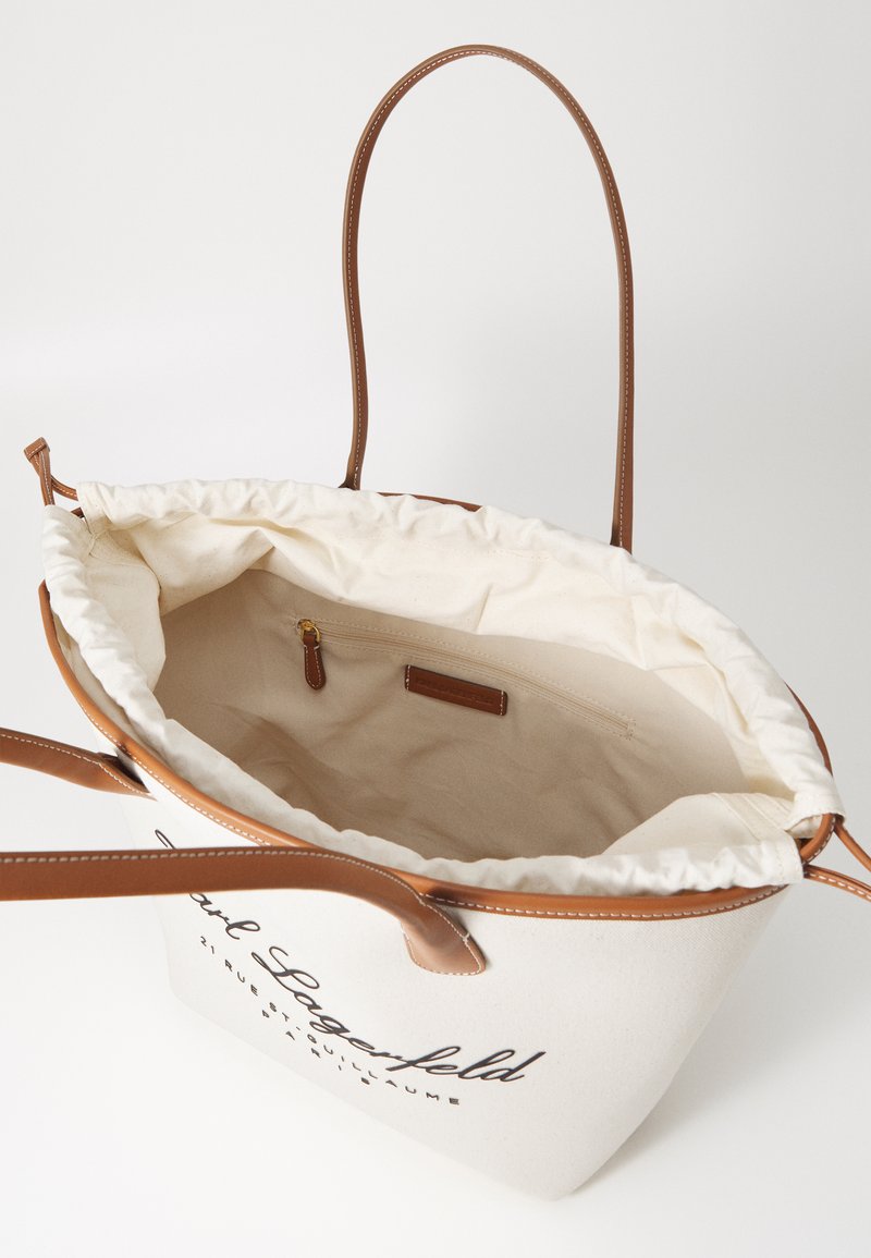 Sac fourre-tout en toile couleur crème avec des accents en cuir marron. Comprend un cordon de serrage et une poche zippée à l'intérieur.