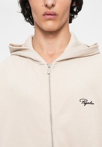 Mladý človek v béžovej rebrovanej mikine na zips s malým čiernym logom "Pegador" na hrudi, s uzavretými perami a neutrálnym výrazom.