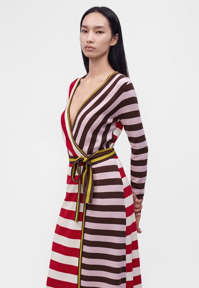 Femme aux longs cheveux noirs portant une robe portefeuille rayée aux rayures horizontales rouges, marron, roses et blanches, avec une ceinture jaune nouée à la taille.