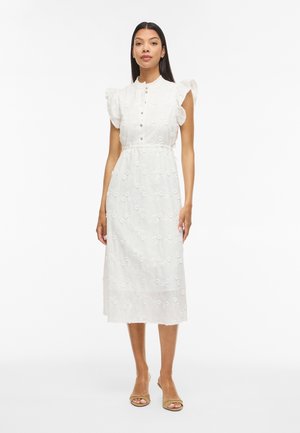 Femme portant une robe midi blanche avec broderies florales, manches ballon à volants, devant boutonné et talons ouverts beiges, debout sur un fond blanc.
