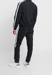 Svart idrottsjacka och byxor med vita randiga detaljer. Jackan har en hög krage och elastisk midja. Skorna är vita med grå detaljer.