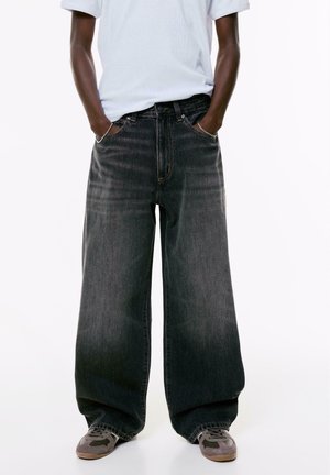 Persoon in te grote vervaagde zwarte jeans, wit T-shirt ingestopt, met handen in de zakken en grijze sneakers op een witte achtergrond.