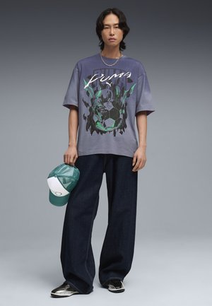 Giovane uomo che indossa una T-shirt oversize viola con grafica Puma, jeans a gamba larga scuri, sneakers bianche e nere, e tiene un cappellino verde e bianco nella mano sinistra.
