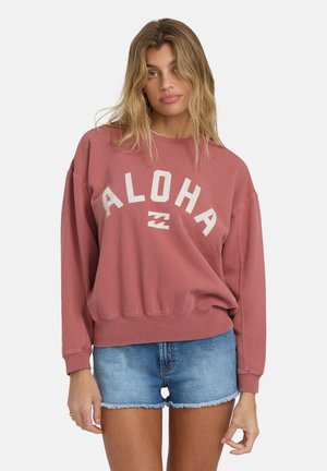 Sudadera rosa con la palabra "ALOHA" en letras blancas, corte relajado, puños y dobladillo acanalados. Combinada con shorts de mezclilla azules con bordes deshilachados.