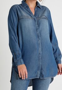 Chemise en denim d’un bleu moyen, dotée d’une fermeture à boutons, de manches longues et d’un ourlet légèrement arrondi. Coutures légères en contraste.