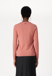 Langärmliger Pullover aus korallenrosa Strickstoff, mit Rundhalsausschnitt und dezentem Logo-Detail auf dem Rücken. Trägt einen schwarzen Rock dazu.