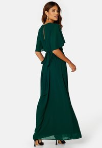 Robe longue verte en tissu léger, avec des manches volantes, une taille nouée et un détail de dos en forme de trou de serrure. Portée avec des talons noirs.