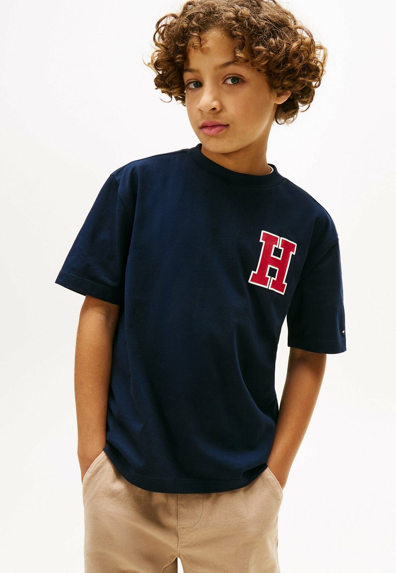 T-shirt in cotone blu navy con logo rosso "H" sul petto, maniche corte e scollatura rotonda classica. Abbinato a pantaloni beige.