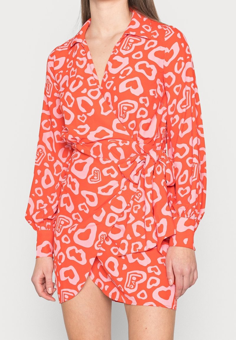 Femme portant une robe portefeuille orange vif avec des manches ballon et un motif cœur rose et "love", nouée à la taille.