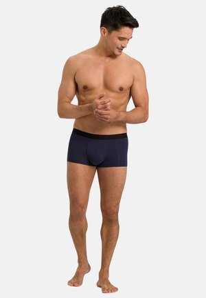 Uomo senza maglietta con capelli corti e scuri che indossa boxer blu navy con elastico nero, in piedi a piedi nudi su uno sfondo chiaro, che guarda verso il basso.